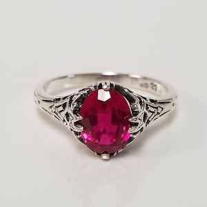 May include: A silver ring with an oval-shaped, bright red gemstone set in a filigree design.