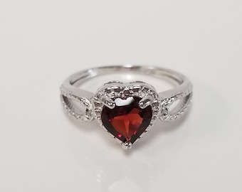Op de afbeelding: Een zilveren ring met een hartvormige rode edelsteen omringd door kleinere heldere edelstenen.