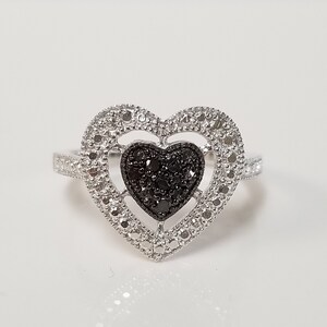 May include: A silver ring with a black diamond heart in the center. The heart is surrounded by a silver heart-shaped frame with smaller diamonds.