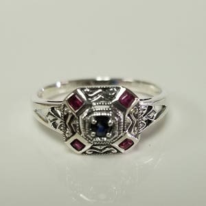 May include: A silver ring with a blue sapphire center stone and four red ruby accent stones. The ring has an intricate design with filigree details.