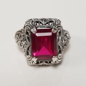 May include: A silver ring with an ornate filigree design and a large, bright red emerald-cut gemstone.