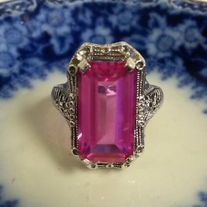 May include: A silver ring featuring a large, rectangular, bright pink gemstone. The ring has an ornate, filigree design around the stone and on the band. The ring is set on a white and blue patterned surface.