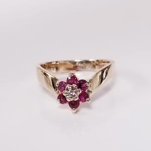 Op de afbeelding: Een gouden ring met een bloemvormige zetting. Het midden van de bloem is een heldere edelsteen omringd door zes rondgeslepen dieproze robijnen. De ringband is glad en gepolijst en loopt iets taps toe naar de centrale edelsteengroep.
