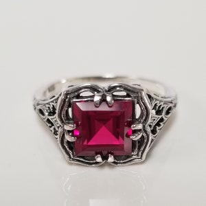 May include: A sterling silver ring with a square, deep red gemstone. The ring features an ornate, filigree design around the gemstone and on the band. The gemstone is the focal point, set in a decorative setting.