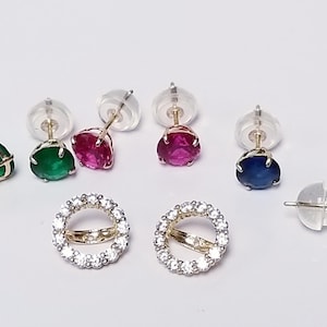 May include: A collection of stud earrings featuring round-cut gemstones in green, red, and blue, alongside a pair of circular earrings encrusted with small clear stones. Each earring includes a metal post and a clear silicone backing for secure wear.