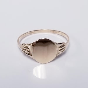 May include: A gold signet ring with a polished, oval face. The ring has a simple band and decorative shoulders. The ring is shown on a white surface.