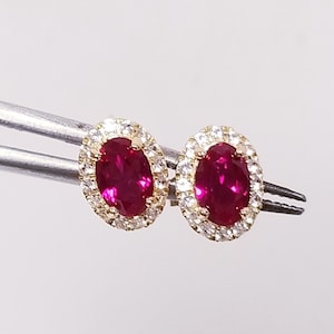 May include: A pair of oval-shaped ruby stud earrings. Each earring features a vibrant red ruby surrounded by a halo of small, clear gemstones. The earrings are set in a gold-colored metal.