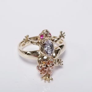 May include: A gold-toned ring featuring a frog design with ruby eyes and a diamond-studded belly. The frog has outstretched arms and is adorned with a tri-color bow. The ring is a unique piece of jewelry.
