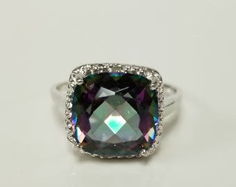 Op de afbeelding: Een zilveren ring met een grote, vierkante, iriserende edelsteen omringd door een halo van kleinere, witte edelstenen.