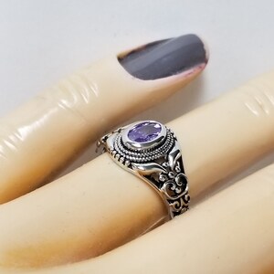 May include: A silver ring with an oval-shaped purple gemstone set in a filigree design.
