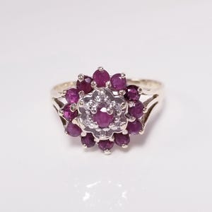 Op de afbeelding: Een goudkleurige ring met een bloemmotief. Het midden van de ring is versierd met kleine, heldere stenen, omringd door een cluster van dieprode edelstenen. De band heeft een gespleten ontwerp aan weerszijden van het bloemvormige middelpunt.