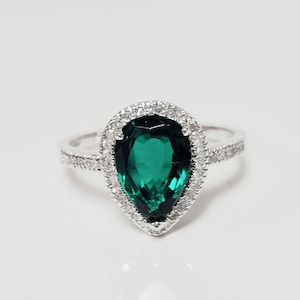 May include: A pear-shaped emerald green gemstone ring with a halo of smaller clear gemstones.
