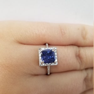 Op de afbeelding: Een zilveren ring met een vierkante blauwe edelsteen omringd door kleinere witte edelstenen.