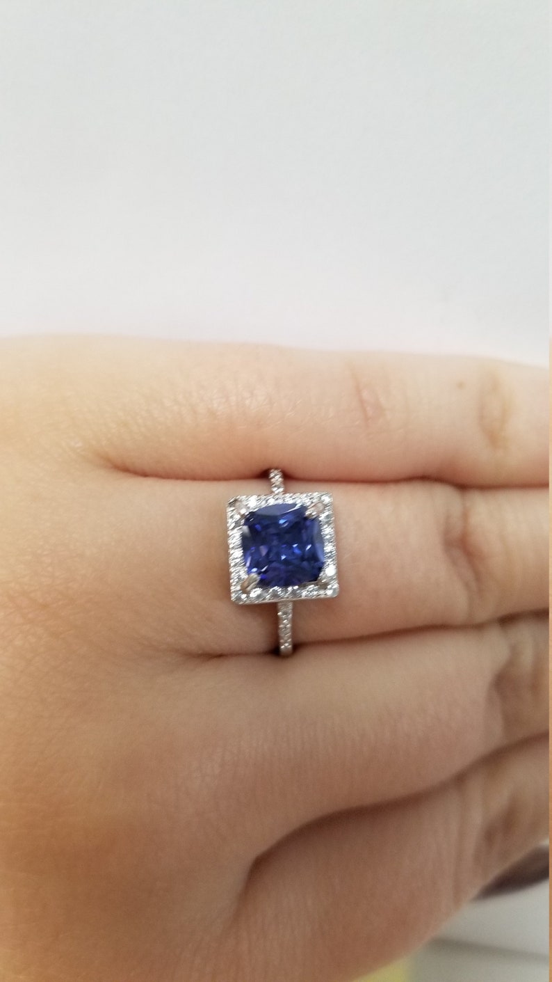 Op de afbeelding: Een zilveren ring met een vierkante blauwe edelsteen omringd door kleinere witte edelstenen.