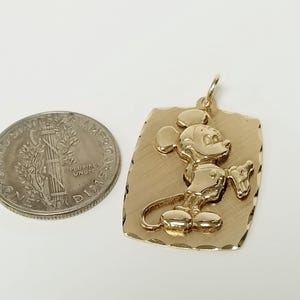 K&ouml;nnte beinhalten: Goldfarbener Anh&auml;nger mit erhabenem Bild von Mickey Mouse auf strukturiertem rechteckigem Hintergrund. Eine US-amerikanische 10-Cent-M&uuml;nze dient zum Gr&ouml;&szlig;envergleich. Der Anh&auml;nger hat eine Schlaufe f&uuml;r eine Kette.