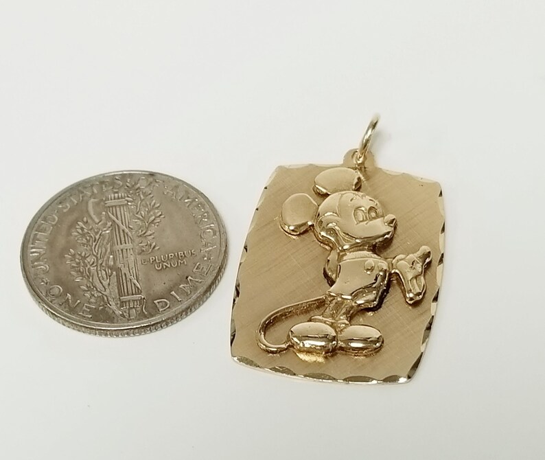K&ouml;nnte beinhalten: Goldfarbener Anh&auml;nger mit erhabenem Bild von Mickey Mouse auf strukturiertem rechteckigem Hintergrund. Eine US-amerikanische 10-Cent-M&uuml;nze dient zum Gr&ouml;&szlig;envergleich. Der Anh&auml;nger hat eine Schlaufe f&uuml;r eine Kette.