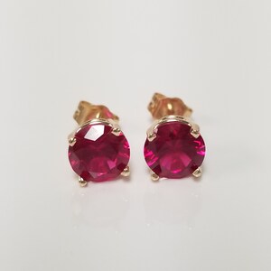 May include: A pair of gold stud earrings, each featuring a large, round, vibrant red gemstone. The gemstones are set in a classic four-prong setting, showcasing their color and brilliance. The earrings are displayed against a plain white background.