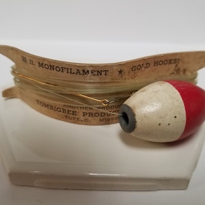 May include: A wooden spool of 20 ft. monofilament fishing line with gold hooks. The spool is labeled "Tombigbee Products, Tupelo, Miss.". A white and red bobber is attached to the line.