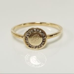 May include: A gold ring with a round, bezel-set centre stone. The centre stone is surrounded by smaller diamonds. The ring is engraved with the letter "P".