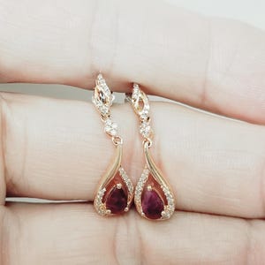 May include: Rose gold teardrop earrings with a deep red gemstone center and a halo of small, clear crystals. The earrings have a delicate, ornate design with a post-back closure. The gemstones are pear-shaped.