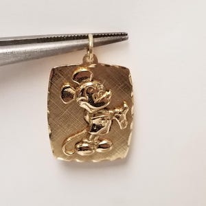 K&ouml;nnte beinhalten: Goldfarbener, rechteckiger Anh&auml;nger mit erhabenem Bild von Mickey Mouse. Der Anh&auml;nger wird von einer Pinzette gehalten. Der Hintergrund hat ein strukturiertes, geb&uuml;rstetes Aussehen. Wahrscheinlich f&uuml;r eine Halskette oder ein Armband.