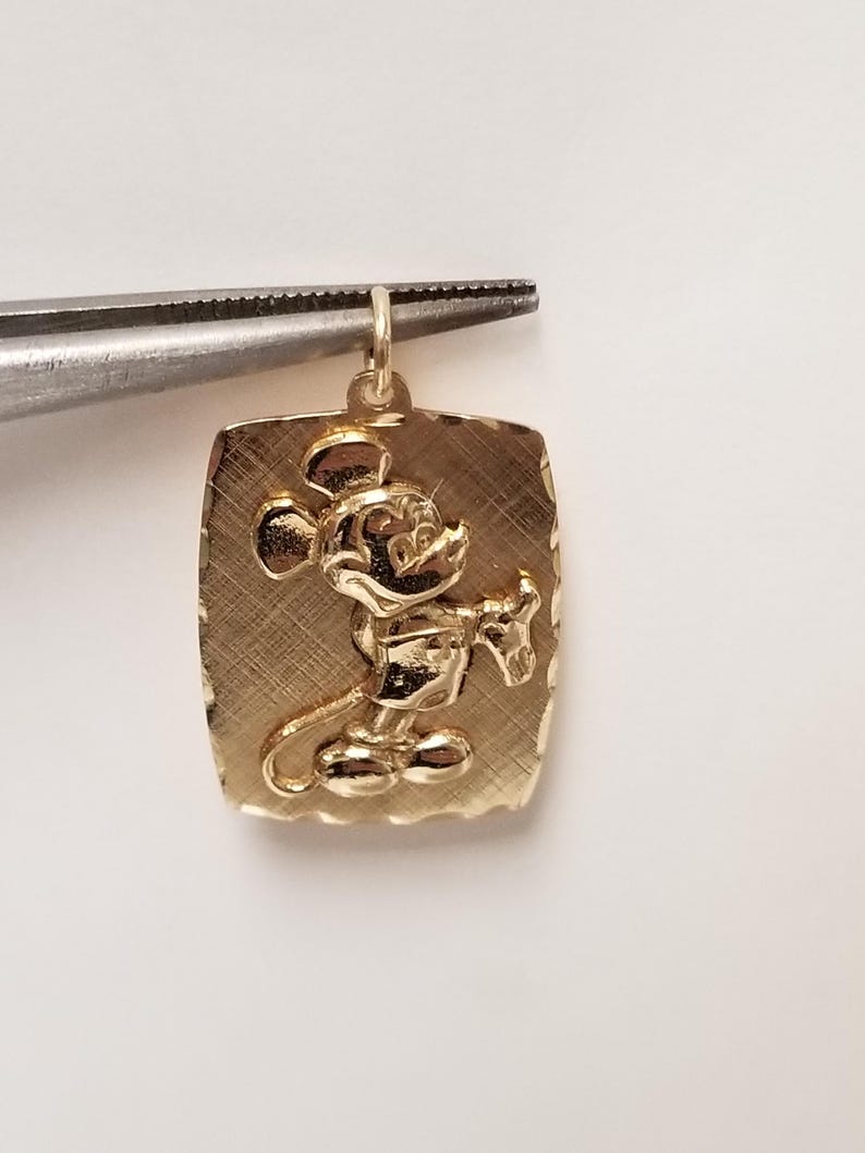 K&ouml;nnte beinhalten: Goldfarbener, rechteckiger Anh&auml;nger mit erhabenem Bild von Mickey Mouse. Der Anh&auml;nger wird von einer Pinzette gehalten. Der Hintergrund hat ein strukturiertes, geb&uuml;rstetes Aussehen. Wahrscheinlich f&uuml;r eine Halskette oder ein Armband.