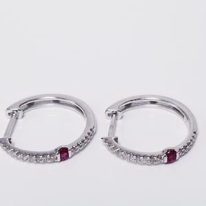May include: A pair of silver-toned hoop earrings featuring a row of small, clear pavé-set stones along the outer edge, accented by a single, round, deep red ruby gemstone set in the center of each hoop. The earrings have a hinged post closure.