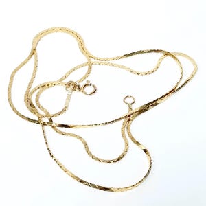 May include: A thin gold chain necklace featuring a flat, textured herringbone-style link design. The necklace includes a small circular spring ring clasp and a matching jump ring at the ends, creating a continuous, reflective metallic appearance.