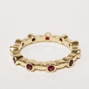 May include: A yellow gold eternity band featuring a series of small, round, bezel-set red rubies spaced evenly around the entire circumference of the ring. The metal has a polished, reflective finish, and the stones are set in individual gold settings.