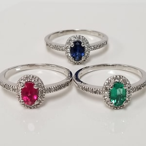 May include: Three silver rings with oval-shaped gemstones set in a halo of smaller diamonds. The rings feature a blue sapphire, a red ruby, and a green emerald.