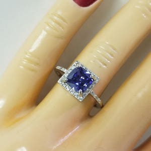 Op de afbeelding: Een vierkante verlovingsring met een grote, donkerpaarse edelsteen omringd door kleinere, heldere edelstenen. De ring is gezet in een zilveren band.