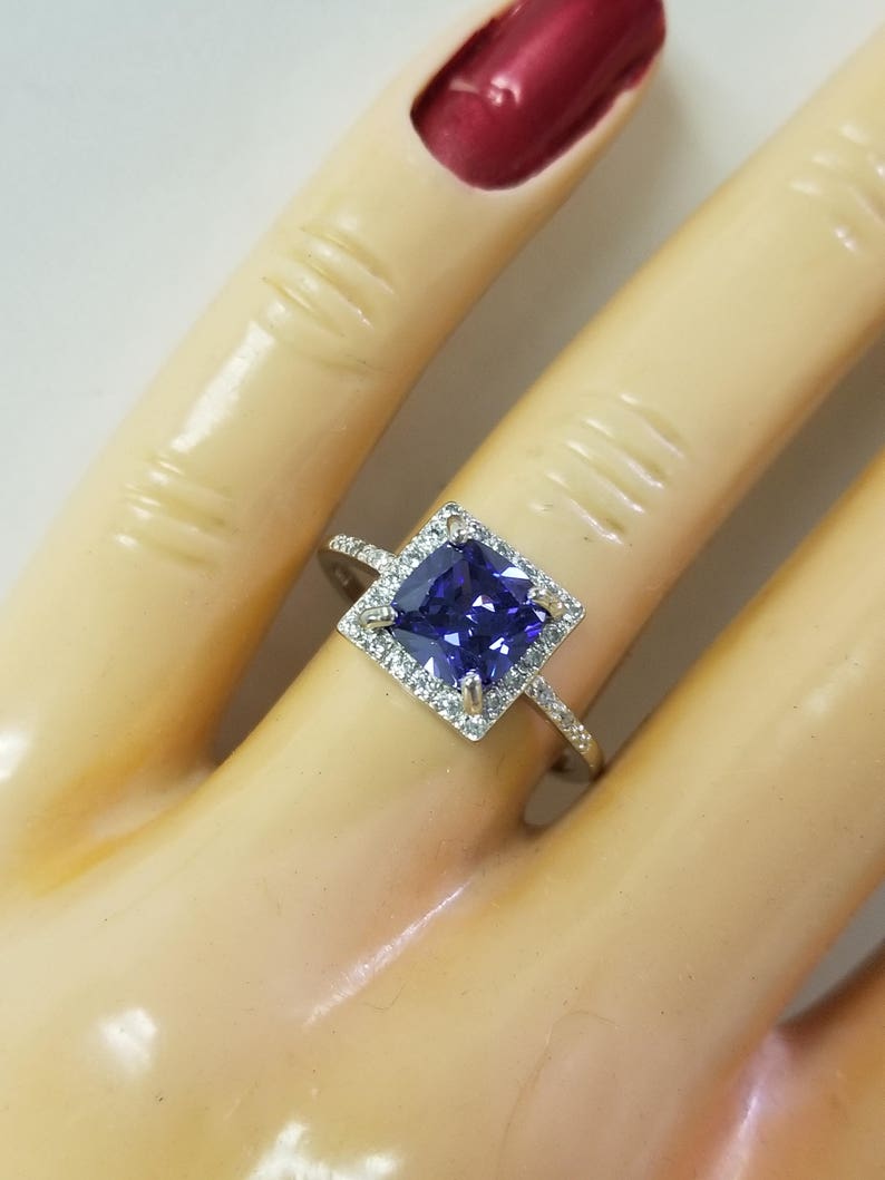 Op de afbeelding: Een vierkante verlovingsring met een grote, donkerpaarse edelsteen omringd door kleinere, heldere edelstenen. De ring is gezet in een zilveren band.