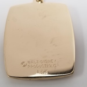 K&ouml;nnte beinhalten: Goldfarbener Anh&auml;nger mit rechteckiger Form und abgerundeten Ecken. Der Anh&auml;nger tr&auml;gt die Gravur "WALT DISNEY PRODUCTIONS" und "14K". Der Anh&auml;nger ist an einer goldfarbenen Kette befestigt.
