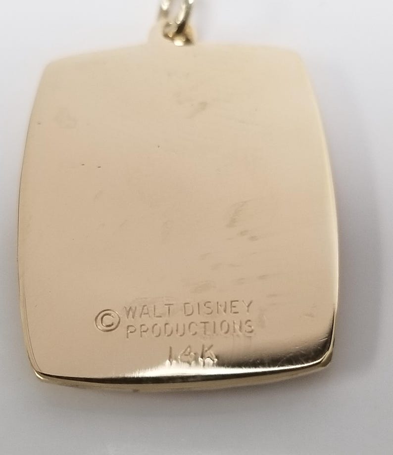K&ouml;nnte beinhalten: Goldfarbener Anh&auml;nger mit rechteckiger Form und abgerundeten Ecken. Der Anh&auml;nger tr&auml;gt die Gravur "WALT DISNEY PRODUCTIONS" und "14K". Der Anh&auml;nger ist an einer goldfarbenen Kette befestigt.