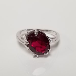 May include: A silver ring with a large, oval-shaped, dark red gemstone. The ring has a delicate, twisted band.