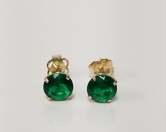 May include: A pair of gold stud earrings featuring round, vivid green gemstones. The earrings are set in a classic four-prong setting, highlighting the bright green colour of the stones.