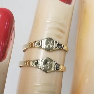 May include: Two gold rings, each featuring a hexagonal center with an engraved initial. The rings have decorative swirls on the sides. One ring has the letter "A", and the other has the letter "S".