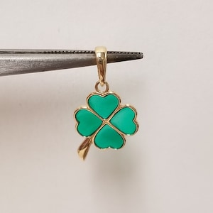 May include: A gold-colored pendant in the shape of a four-leaf clover, with each leaf filled with a vibrant green color. The pendant is held by a pair of tweezers against a white background. The clover design is a symbol of luck.