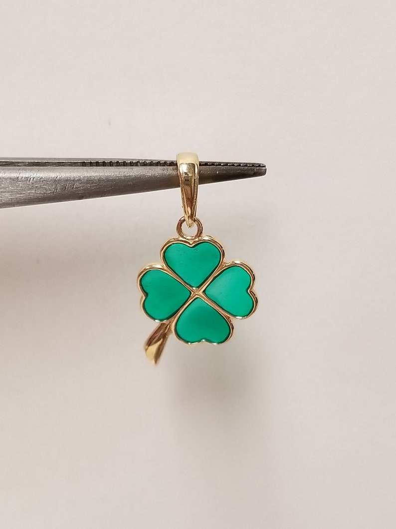 May include: A gold-colored pendant in the shape of a four-leaf clover, with each leaf filled with a vibrant green color. The pendant is held by a pair of tweezers against a white background. The clover design is a symbol of luck.