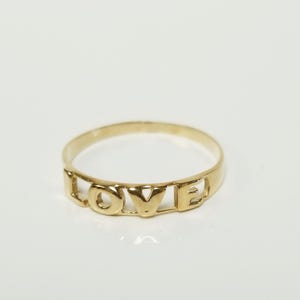 May include: A gold ring with the word "LOVE" spelled out in capital letters. The ring is a simple band design with the letters cut out, creating a delicate and stylish piece of jewelry.