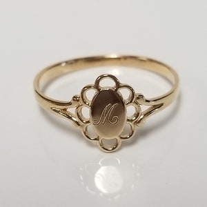 May include: A gold ring with a delicate floral design and an oval-shaped center featuring the letter "M" engraved inside.