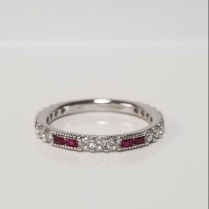 Könnte beinhalten: Ein silberner Bandring mit einer Reihe kleiner klarer Steine und zwei Reihen roter quadratischer Steine.