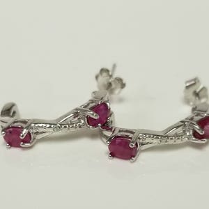 May include: Silver hoop earrings with three ruby gemstones. The earrings are a delicate design with a curved shape.