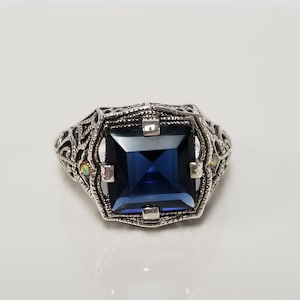 May include: A silver-toned ring featuring a large, square-cut dark blue gemstone held by four prongs. The band displays an intricate, openwork filigree pattern with small, iridescent accent stones embedded on the sides of the setting.
