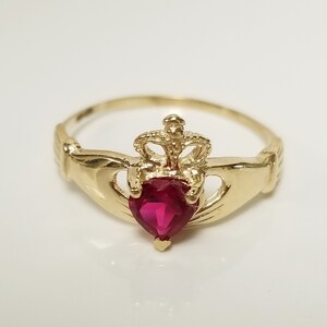 May include: A gold ring with a red heart-shaped stone and a crown design. The ring is a Claddagh ring, a traditional Irish symbol of love, friendship, and loyalty.