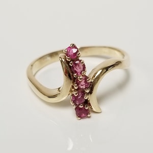 May include: A gold ring featuring a bypass design with a vertical row of five round, faceted pink rubies set in the center. The polished gold band curves around the stones, creating a fluid, open silhouette that highlights the vibrant gemstones.