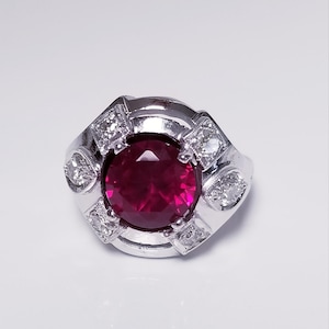 Peut inclure: Une bague en argent avec une grande pierre de rubis rouge ronde entourée de petits diamants clairs.