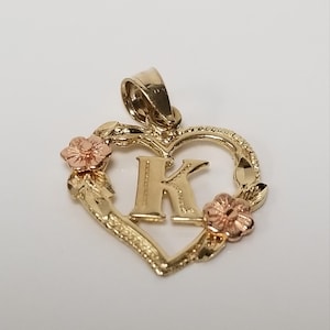 May include: A gold-coloured pendant shaped like a heart, with the letter "K" at its centre. The heart is decorated with two small, rose-gold flowers and leaves. The pendant has a textured edge and a loop for a chain.