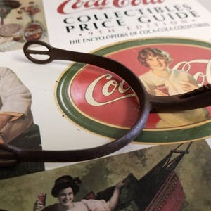 Op de afbeelding: Een roestige bruine metalen sporen met een afgeronde lus aan de bovenkant en een puntig uiteinde. De sporen ligt op een Coca-Cola-boek met een rood en groen Coca-Cola-logo.