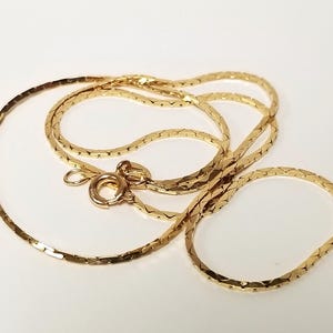 May include: A gold-toned chain necklace featuring a flat, interlocking link design with a reflective surface. The necklace includes a circular spring ring clasp mechanism and a small jump ring, showcasing a continuous, polished metallic finish throughout.